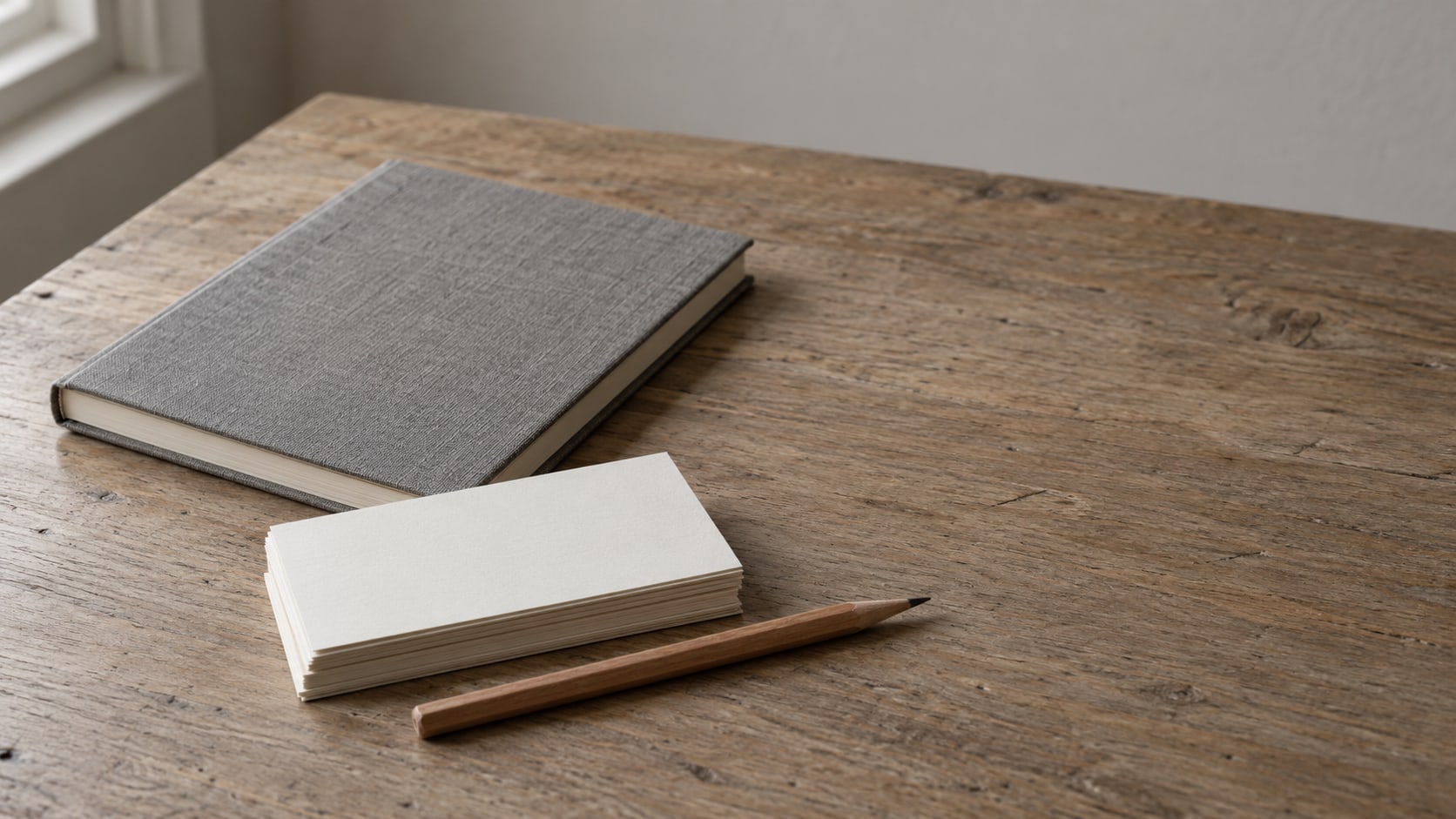 A neat stack of blank index cards beside a sharpened pencil and a closed notebook on a plain wooden desk.