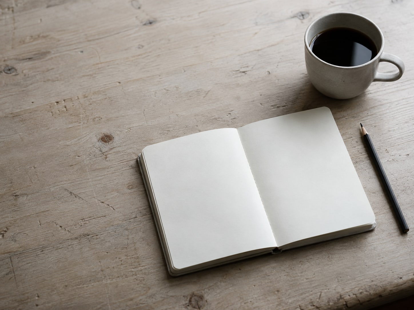 A blank notebook open on a wooden desk beside a sharpened pencil and a cup of coffee.