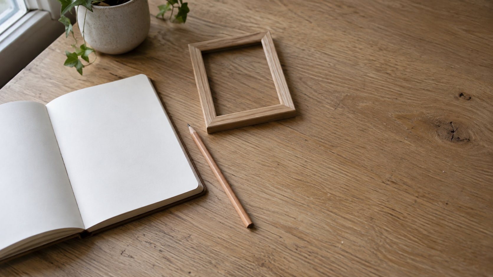 A blank notebook, a sharpened pencil and a small empty wooden frame arranged on a clean oak desk.
