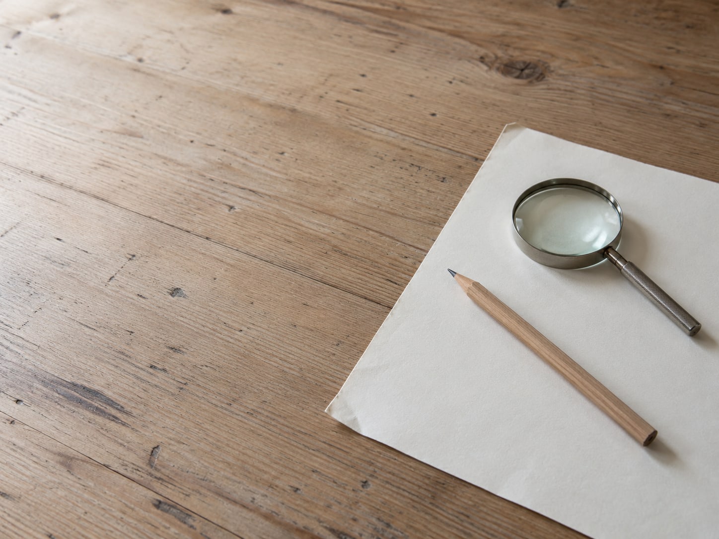 Una hoja de papel en blanco, un lápiz y una lupa sobre un escritorio.
