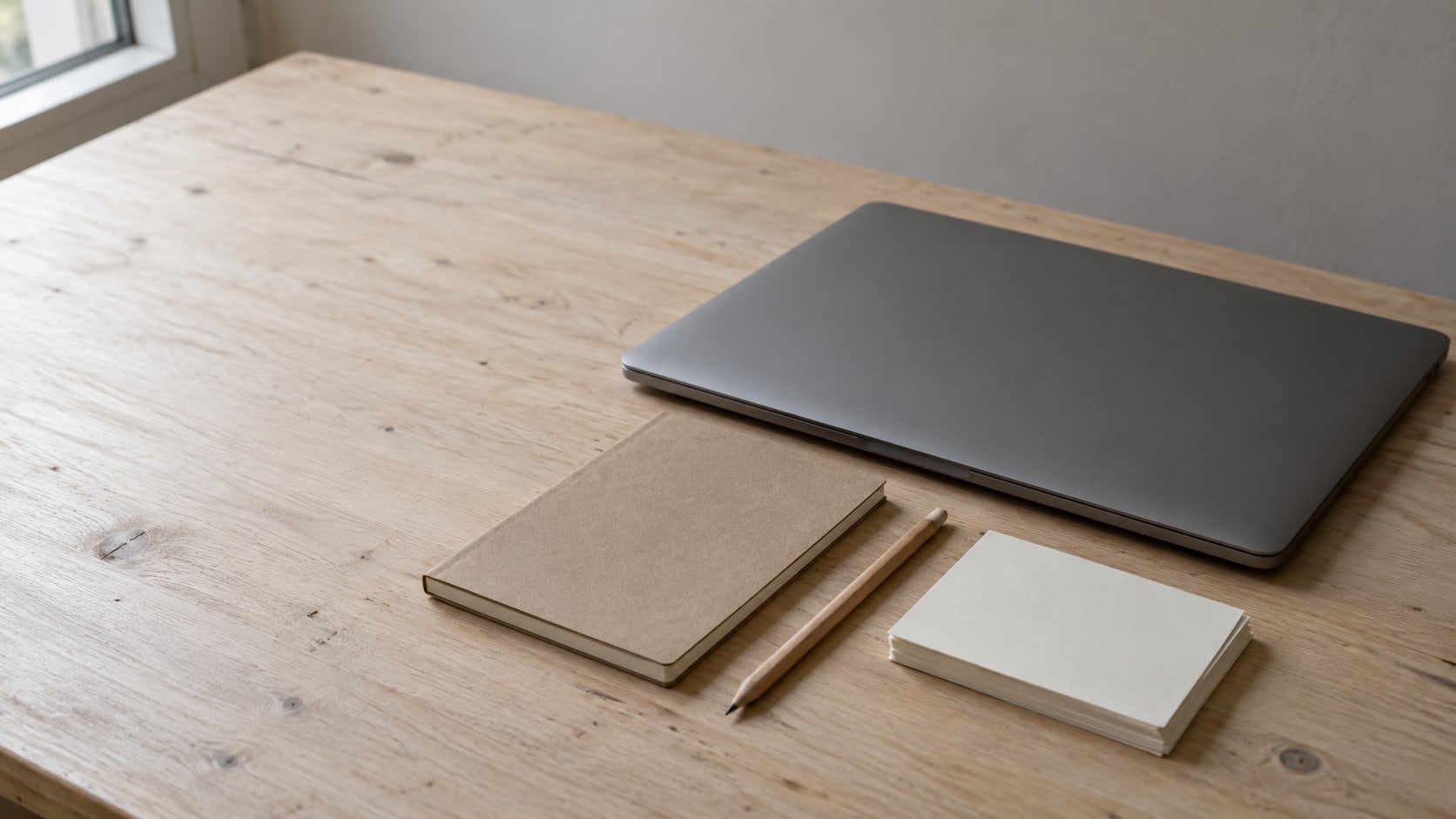 Una computadora portátil cerrada junto a una libreta pequeña, un lápiz y una pila corta de tarjetas en blanco sobre un escritorio de madera clara.