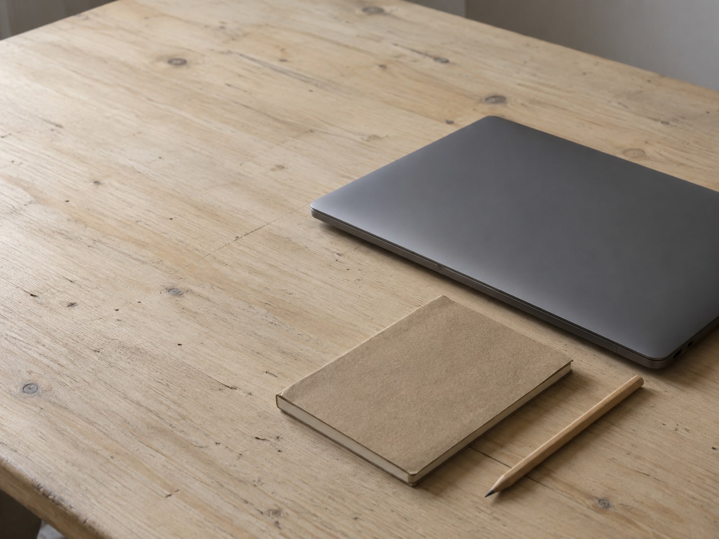 Una computadora portátil cerrada junto a una libreta pequeña y un lápiz sobre un escritorio de madera sencillo.