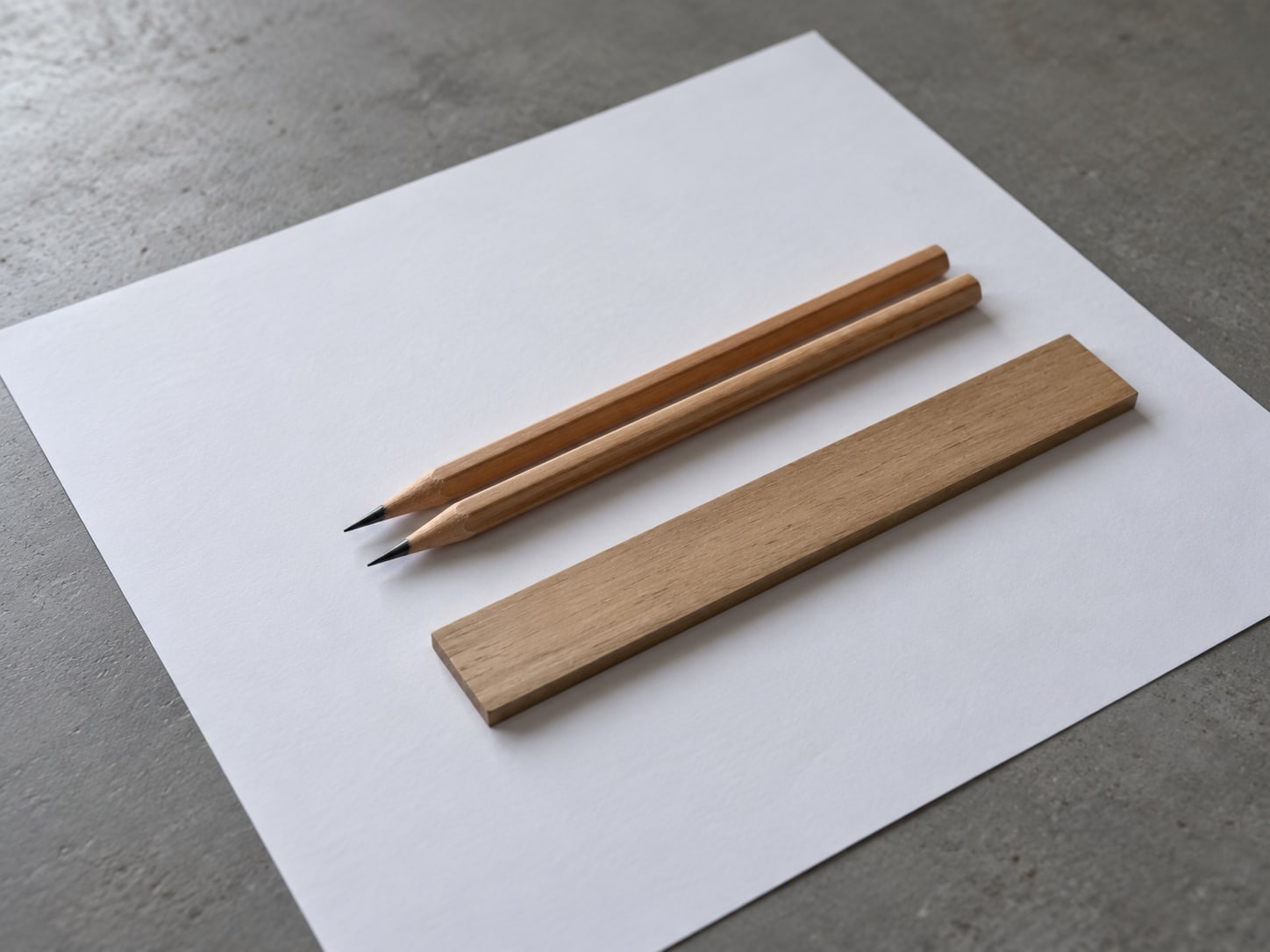Two sharpened pencils lie parallel on a plain sheet of blank paper beside a small wooden ruler.