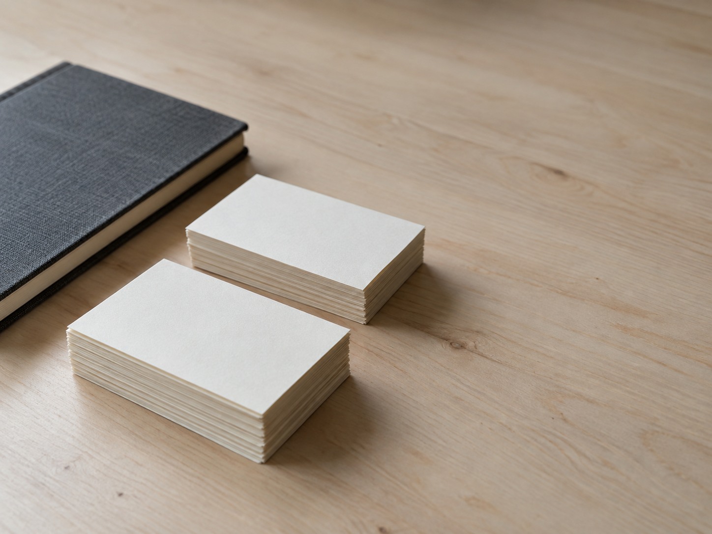 Una pequeña pila de tarjetas índice en blanco dividida en dos montones ordenados sobre un escritorio de madera clara junto a un cuaderno cerrado.