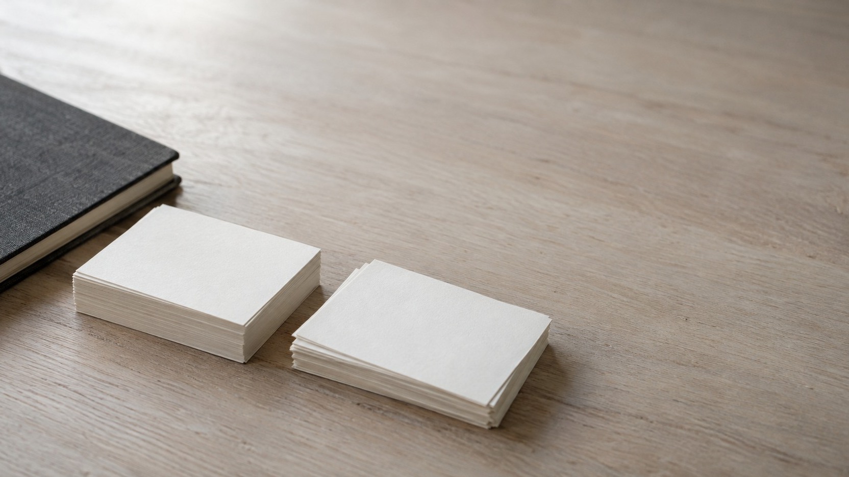 Dos pequeñas pilas de tarjetas índice en blanco, una cuadrada con cuidado y otra puesta un poco aparte, sobre un escritorio de madera clara junto a un cuaderno cerrado.