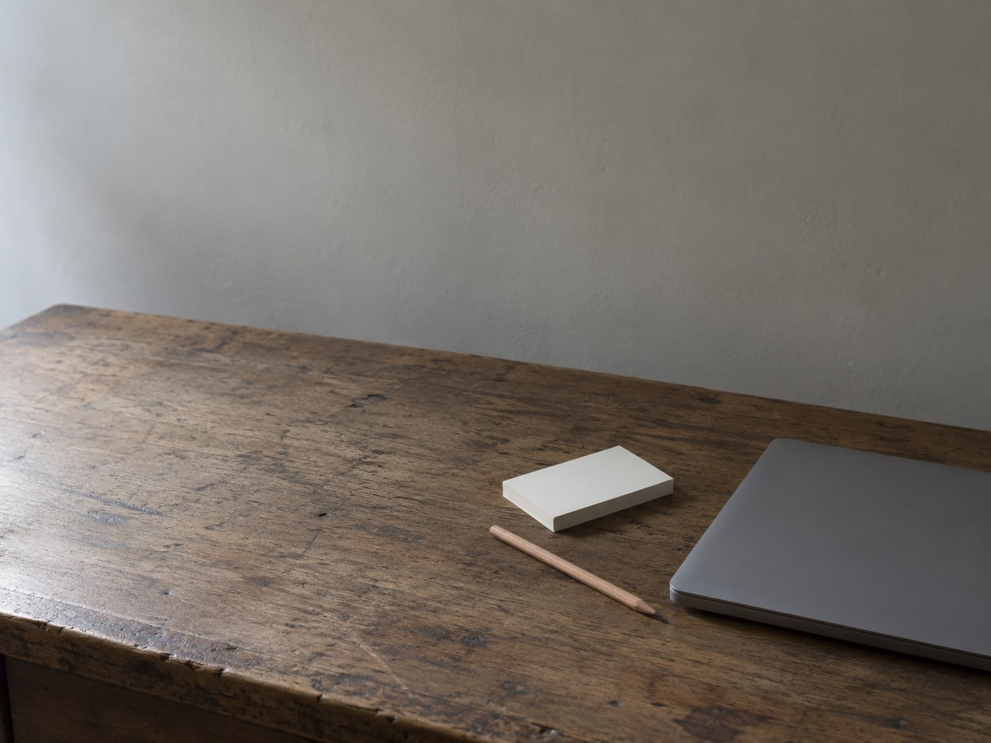 A closed laptop beside a small stack of blank index cards and a pencil on a worn wooden desk.
