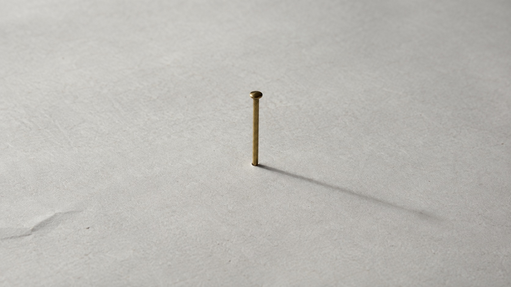 A slender brass pin standing upright on a large sheet of pale paper, casting one long shadow across the empty surface.